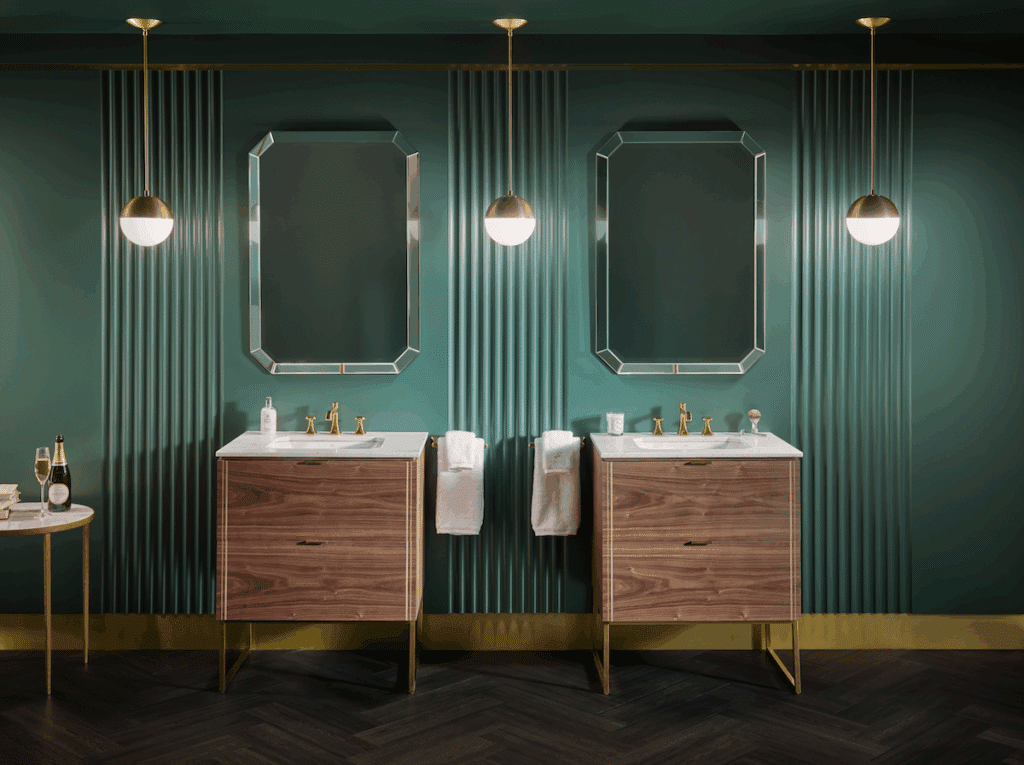 Two mid-century wood vanities with gold faucets against a deep green fluted wall with octagonal mirrors and gold globe pendant lights.