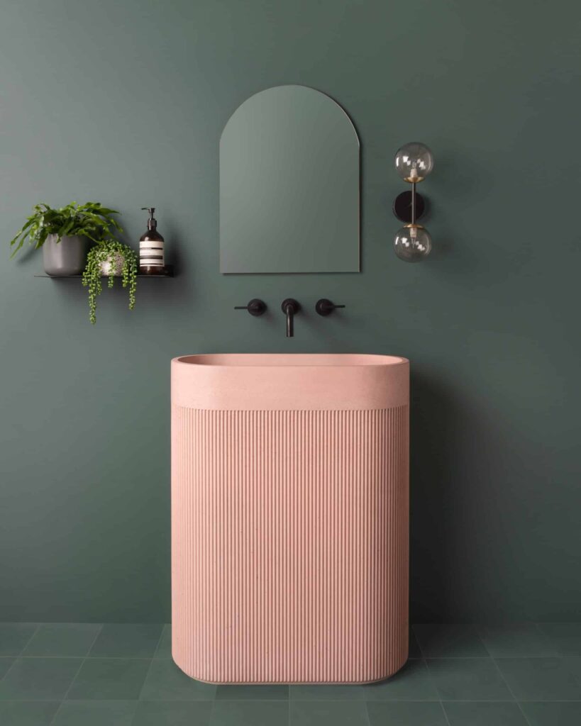 A pink Kast concrete pedestal sink with a fluted texture against a dark green wall with a black wall-mount faucet.