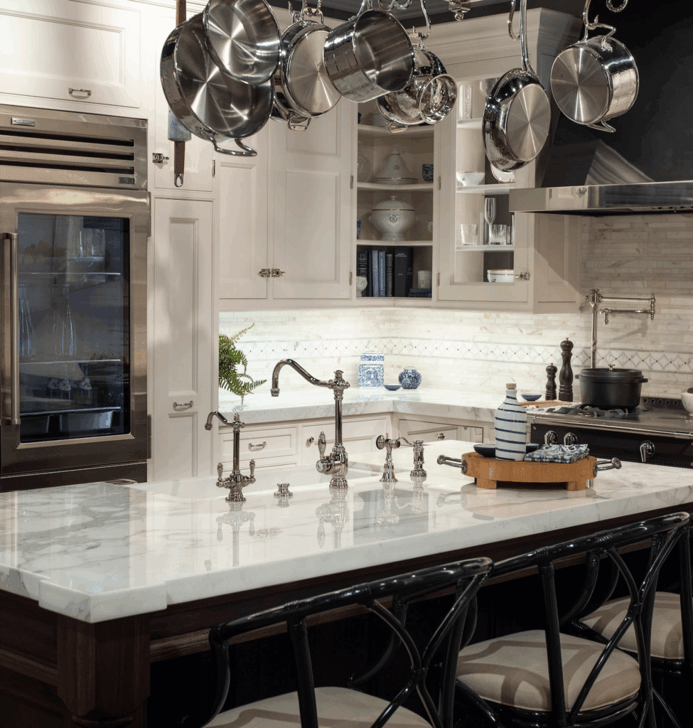 A traditional luxury kitchen with a white marble island, ornate bridge faucet, stainless steel refrigerator, and hanging pot rack.