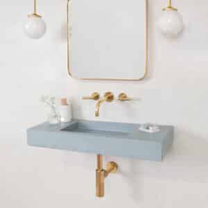 A light blue Kast concrete wall-mounted sink with a gold wall-mount faucet and exposed gold p-trap against a white tile wall.